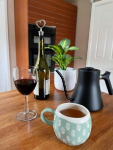 A plant, kettle and cup of tea and a wine bottle and glass of wine all sitting on a kitchen worktop.